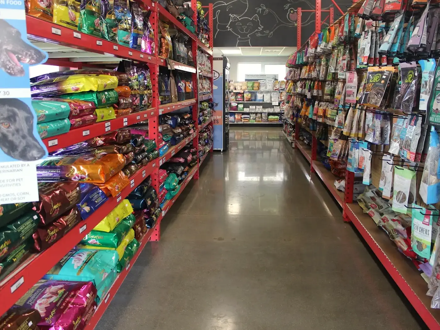 A long aisle of dry dog and cat food bags on red shelving, with treats on the opposite wall